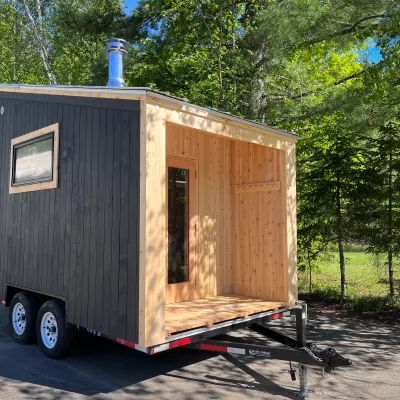 A mobile sauna built on a black dual-axle trailer, featuring dark charcoal vertical siding and a recessed entryway made of natural light wood.