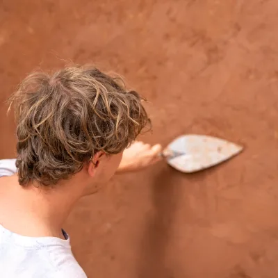 A student is photographed from behind plastering a wall with a trowel.