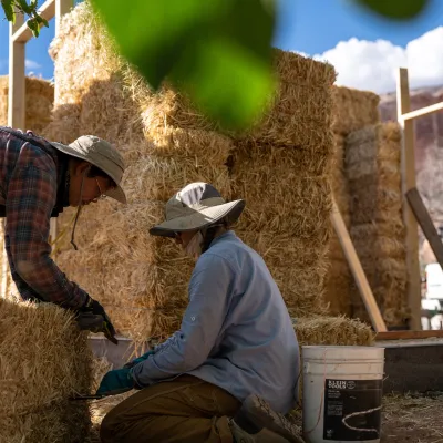 Two people crouch near a strawbale in front of a partially framed building.