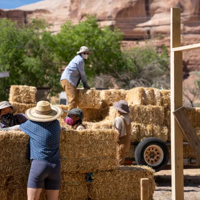 A group 3 students, wearing dust masks, assembling a strawbale wall while 2 students unload additional bales from a trailer in the background. 