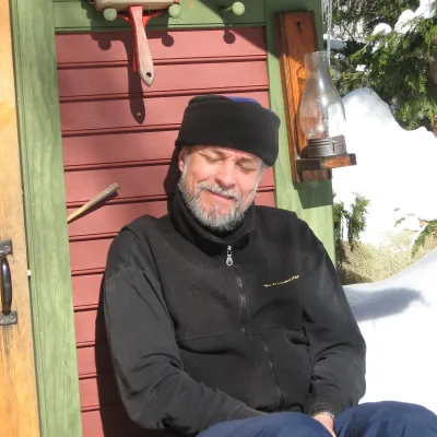 A bearded man in winter clothing sits against the exterior wall of a red-and-green wooden sauna, eyes closed and face turned toward the sun