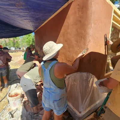 A photo of a job site with many people working. The image centers on three people apply a reddish brown plaster to the corner of a building. 