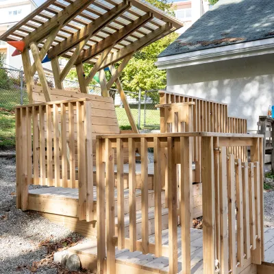 A small wooden structure with railings and a slanted translucent roof stands outdoors on a gravel surface. The design features multiple levels connected by ramps and platforms, blending play and architectural elements. Trees, a fence, and a nearby building are visible in the background under a clear sky.