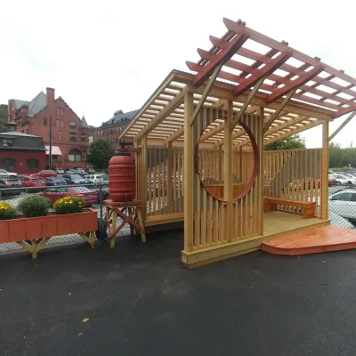 A wooden outdoor pavilion with a slatted roof and circular wall opening stands on a paved surface near a parking lot. The structure includes built-in benches, a ramp for access, and a rain barrel connected to the roof for water collection. A planter box with flowers sits nearby, and historic brick buildings and parked cars are visible in the background.