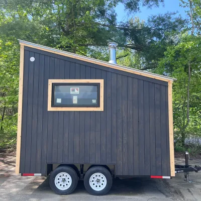 A mobile sauna built on a black dual-axle trailer, featuring dark charcoal vertical siding and a recessed entryway made of natural light wood.