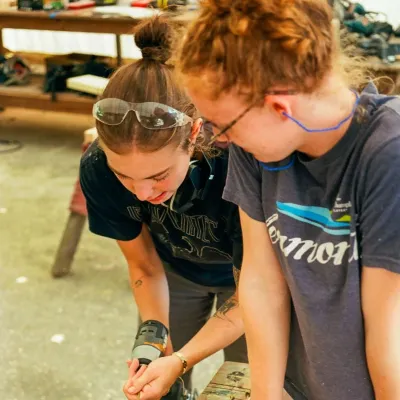 Two students work together on cutting a board, one is holding and the other is preparing to cut with a circular saw.
