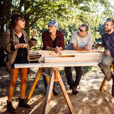 Three students lean on some lumber set on sawhorses while listening to an instructor.