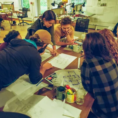A group of students huddles around a table strewn with design drawings in the main studio