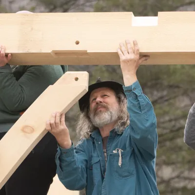 Skip looks up at a timber brace during a raising. He is kneeling and wearing a blue shirt and brimmed hat.