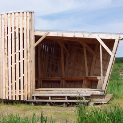 A tall, cylindrical wall of vertical wooden slats wraps around a sheltered wooden platform in a grassy field with mountains in the distance.