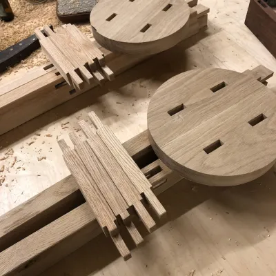 Two stool tops are pictured unassembled next to their legs. The stool top has holes for through-tenon joinery.