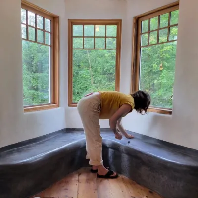 A person is pictured in front of a bay of windows with a bench nook. The person is leaning over and touching the surface of the bench.