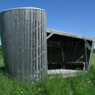 A weather wooden bus stop featuring a unique heliciform shape in a grassy field in front of a blue sky.