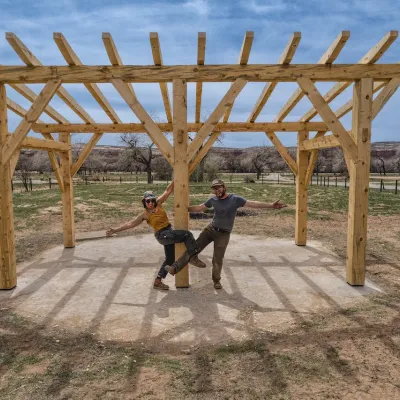 A timber frame pavilion with two people posing with their arms outstretched to showcase the project. ​ 