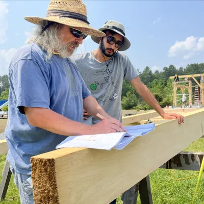 A student and instructor look at plans on a beam in the foreground, with a timber frame in progress in the background.