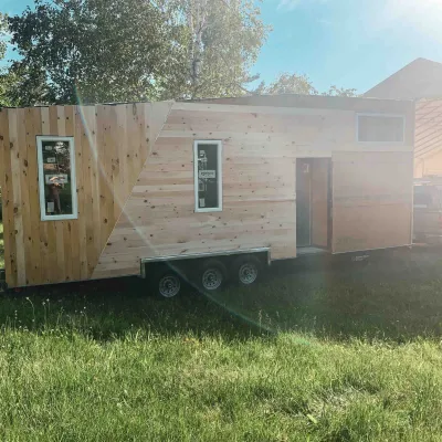 A finished tiny house on a trailer is pictured with wooden siding on a lawn.