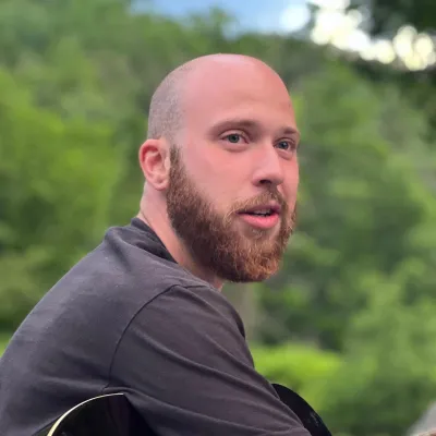 Tyler is photographed outside with trees in the background. He is wearing a gray t-shirt and holding a guitar.