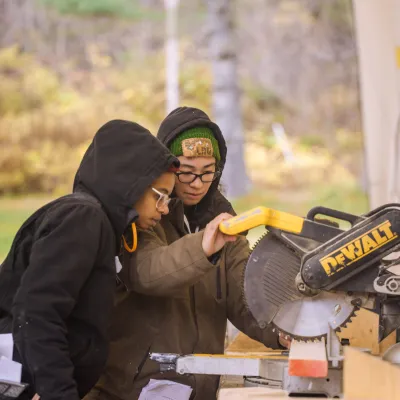 An instructor guides a student at a chop saw station in an outdoor classroom.