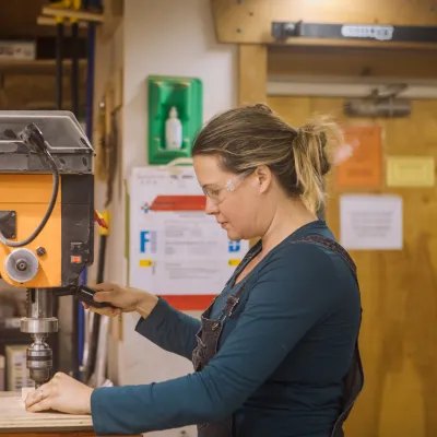 A student with blonde hair in a ponytail wearing a turquoise shirt uses the drill press in the woodshop.