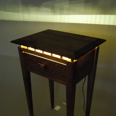 A shaker end table photograped in darkness, with lighting emanating from inside through openings in the drawer.