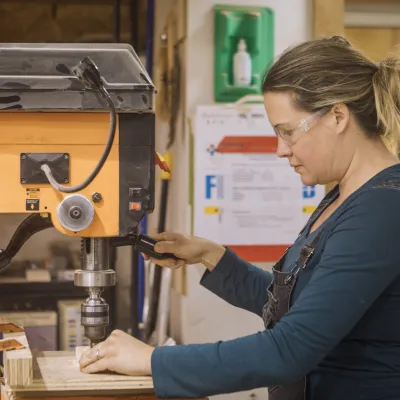 A person wearing a teal shirt uses the drill press in the woodshop.