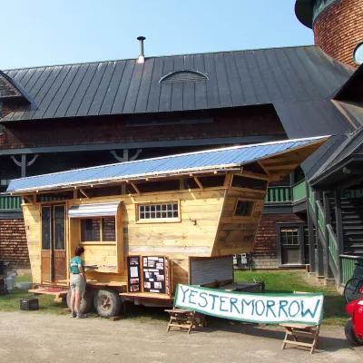 A small wooden tiny house on wheels is displayed outdoors in front of a large rustic building with dark shingles and green trim. Several people are gathered around, and a banner in front of the tiny house reads “Yestermorrow.”