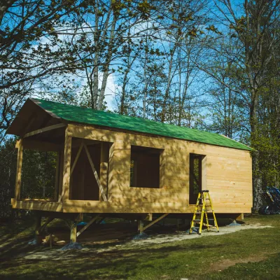 Wide shot of a cabin in the forest, with green roof and wood siding.
